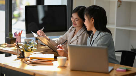 Asian female employees discussing ideas of project looking details on binder folder. Friendly millennial businesswoman talking to female colleague, sitting at table in office.の写真素材