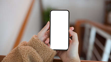 A young female in comfort sweater using a mobile phone to search some details on the internet, close-up female hands holding smartphone blank screen.の写真素材