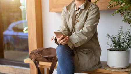 A female in trench coat sitting outside the coffee shop, using her smartphone and waiting public transportation.の写真素材