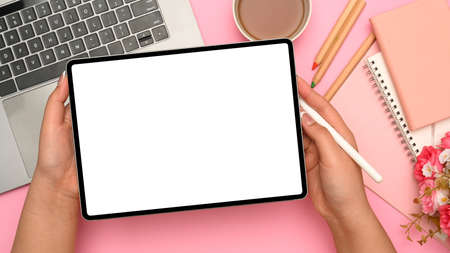 Woman hands with digital tablet touchpad white screen mockup on pink pastel background. workspace with laptop, stationery, teacup and flowers bouquet. top viewの写真素材