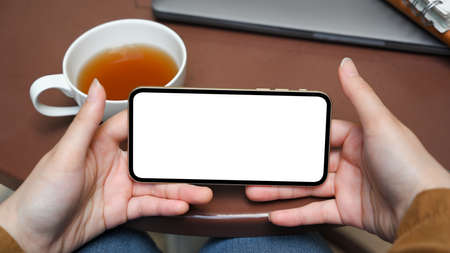Close-up image, Female hands holding a smartphone white screen mockup in a horizontal position over...