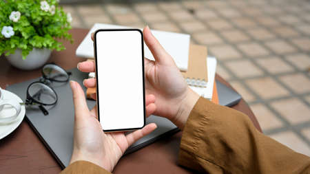 Female hands holding a mobile phone white screen mockup over coffee table in outdoor space. remote...