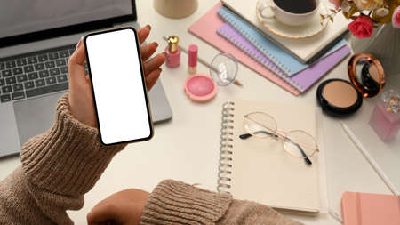 Trendy millennial woman in cozy sweater reading some online articles on her smartphone at her working table. phone white screen mockup.の写真素材
