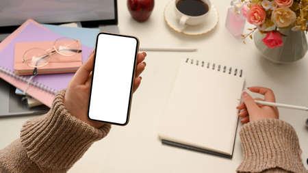 A female freelancer remote working at home, hand writing on empty spiral notepad and holding smartphone white screen mockup over stylish desk.の写真素材