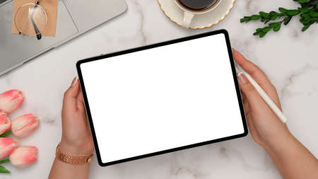Female hands showing tablet white screen mockup over her beautiful office desk. Laptop, coffee cup, glasses, tulips flower and decor on white marble background. top viewの写真素材
