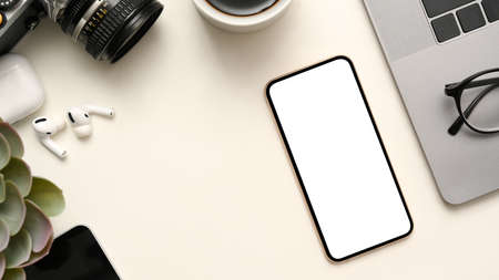 Workspace with camera, decor plant, earbuds, eyeglasses, laptop computer, and mobile phone white screen mockup on white background.の写真素材