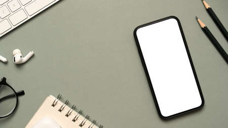 Office workspace table with office essential tools, spiral notepad, earbuds, eyeglasses, pencil, keyboard computer and smartphone mockup on grey background. top viewの写真素材