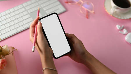 Close-up, Girl holding a cellphone blank screen mockup and pencil over her stylish pink office desk.