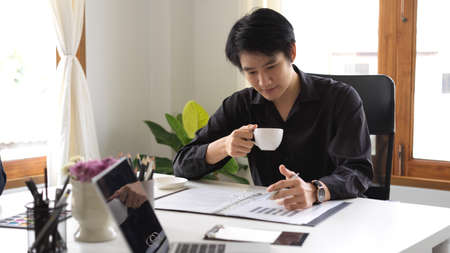 Handsome and smart asian young businessman sipping coffee while looking at the financial data on document folder in his office room.の写真素材