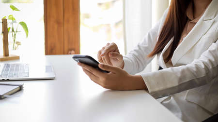 Cropped image, Elegant businesswoman or female manager in white suit using her smartphone at the office desk.の写真素材