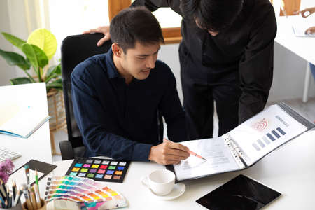 Male graphic designer is looking the information on the document and discussing with his colleague...
