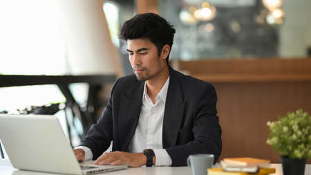 Focused Asian male boss in business suit working on laptop computer in the modern office workplace.