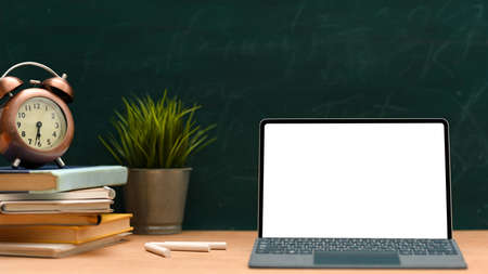 Vintage school table with laptop white screen mockup, stack of books, vintage clock and decor plant on wood table against green chalkboard background.の写真素材