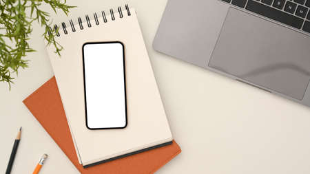 Overhead shot, Smart mobile phone white screen mockup on spiral notepad, pencil laptop, decor plant...