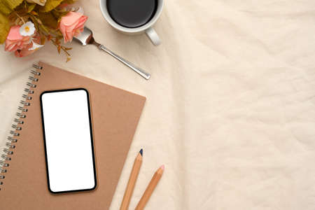 Feminine workspace with smartphone white screen mockup, spiral notebook, a cup of coffee, flowers...