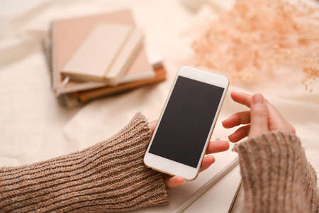 Close-up, Female blogger in warm sweater using smartphone to check her social media at her comfortable working desk. phone black screen mockup.の写真素材