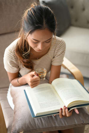 A focused Asian woman concentrates on reading a book while sipping coffee in her living room.