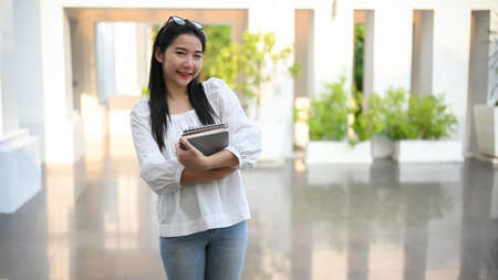 Charming Asian university student standing in front of the campus building and holding her textbook.の写真素材