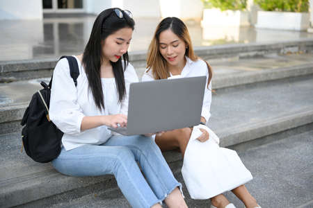 Two Asian female college student sitting on the stairs, working and looking something on laptop computer screen.の写真素材
