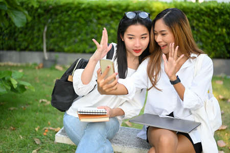 Two attractive asian millennial college women having a video call with her friends on smartphone in a green park.の写真素材