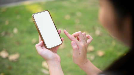Female hands holding a smartphone white screen mockup against blurred green park background. closeup