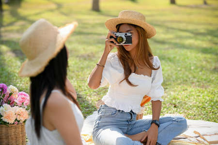 Two female best friends enjoying picnic in the green park together, Female taking a picture of her friends with retro camera.の写真素材