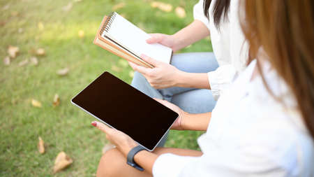 Cropped image, Two female friends sitting in the park together, talking and sharing their ideas, holding a tablet black screen.の写真素材