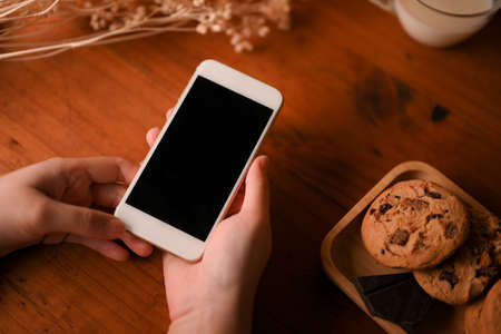 Female holding smartphone black screen mockup over wooden table with a plate of cookies and decor.の写真素材