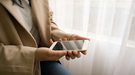 Close-up image, Young woman in casual suit sitting near the window and using holding a smartphone.の写真素材