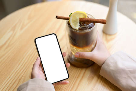 Female relaxes in the coffee shop, holding an iced coffee and smartphone white screen mockup over wood table. Close-up imageの写真素材