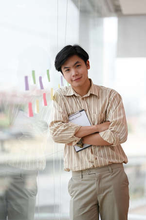 Asian young male worker employee standing near the large window, arms crossed with clipboard paper.