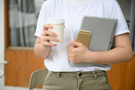 Cropped image, Female college student walking out of the coffee shop with a cup of coffee, a laptop...