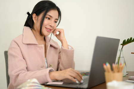 Attractive Asian female graphic designer working on laptop computer in minimalist office workspace.