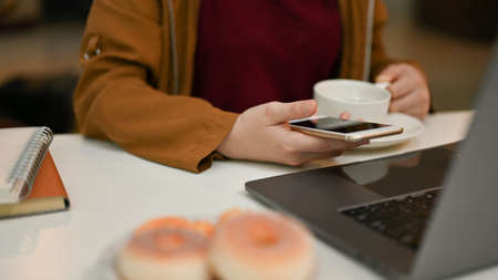 Close-up image, Female freelancer remote working at the coffee shop co-working space, using smartphone and laptop computer while sipping coffee.の写真素材
