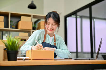 Happy Asian female entrepreneur work on parcel boxes, write a receipts and customer address on the box. SME business owner conceptの写真素材