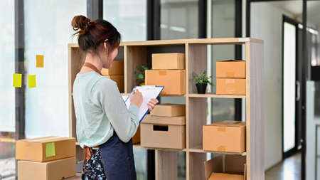 Charming Asian millennial woman fashion retail shop owner checking her clothes and accessories stock...
