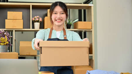Cheerful Asian young female online shop entrepreneur holding a cardboard box or parcel box in the...