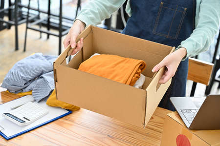 Female fashion clothes shop owner holding a parcel box with folded clothes, preparing to pack and...