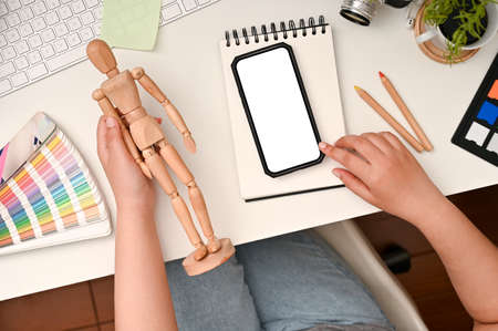 Top view, Female graphic designer sitting at her desk, holding a wood human figure and using smartphone. Phone white screen mockup.の写真素材