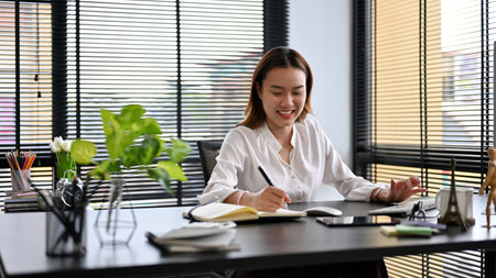 Cheerful Asian female manager sits at her desk in a private office workplace, working and managing...