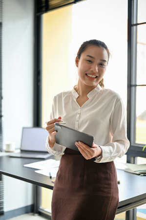 Portrait of a friendly Asian female manager, female boss leaning on her office desk, holding a...