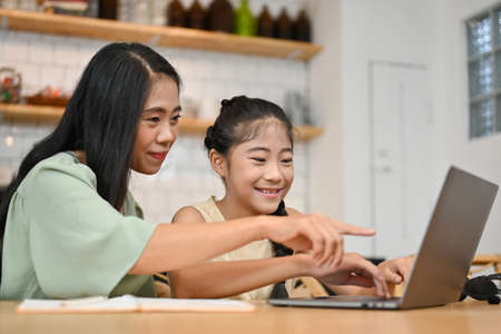 Cheerful Asian mom looking at laptop screen, helping her daughter on an online homework, explaining...