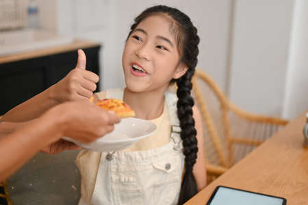 A happy Asian young girl gives her mom a thumbs up after giving her a tasty snack while she was doing homework.の写真素材