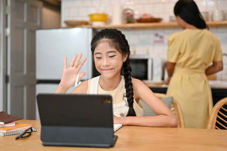 Pretty Asian young girl studying on online class via tablet computer, sits in dining room while her mom is cooking in the kitchen behind.の写真素材