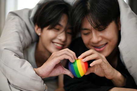 Lovely asian young gay couples lying on bed under the blanket and holding a rainbow LGBT heart...