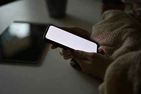 Close-up image, A female hands holding a smartphone white screen mockup, using cellphone at desk in...
