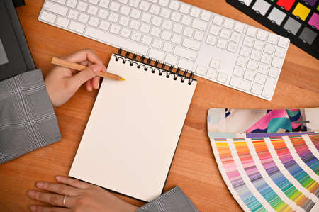 Graphic designer office desk with a female hand writing something on blank spiral notepad. top view...