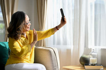Happy retired asian woman sits on the couch, enjoy talking and video calling with her daughter on...