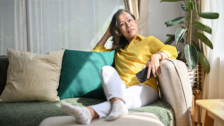 Beautiful Asian middle-aged woman relaxes in the living room, sitting on comfortable sofa and watching TV.の写真素材