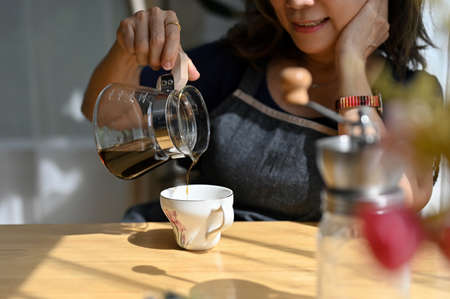 Cropped, close-up image. Beautiful asian aged woman holding a coffee jar, poring hot coffee into a...